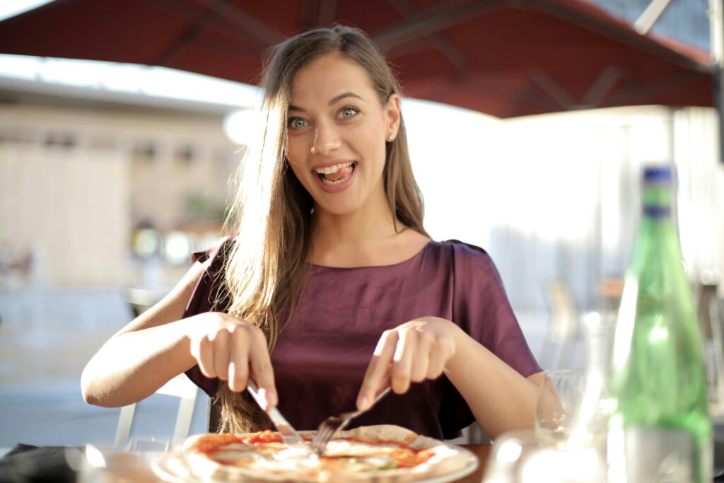 A happy woman wearing a purple top enjoys eating pizza at an outdoor restaurant.